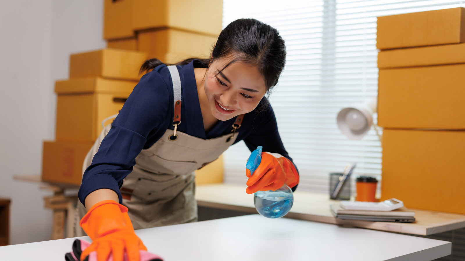A cleaner wiping down a desk in a room stacked with moving boxes.